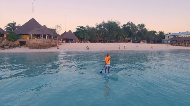 AERIAL: Stand up paddling in the Indian Ocean against the background of an orange sunset near the coast of Africa. African man on SUP board. 4K.