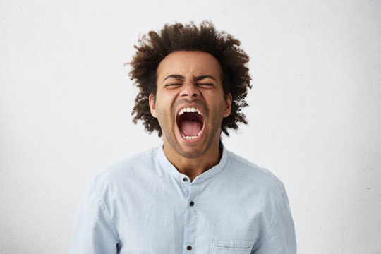 Portrait of dark-skinned African guy with curly hair screaming with wide opened mouth closing his eyes in despair having unhappy expression. Frustrated male shouting loudly having aggression
