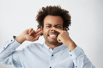 Afro American dark-skinned male frowning his face holding finger on nose and hand on ear trying to...