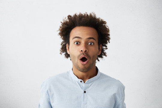Portrait of astonished male model with curly hair, dark eyes and bristle opening his mouth with surprisment hearing terrified news. Excited man in formal clothes isolated over white background