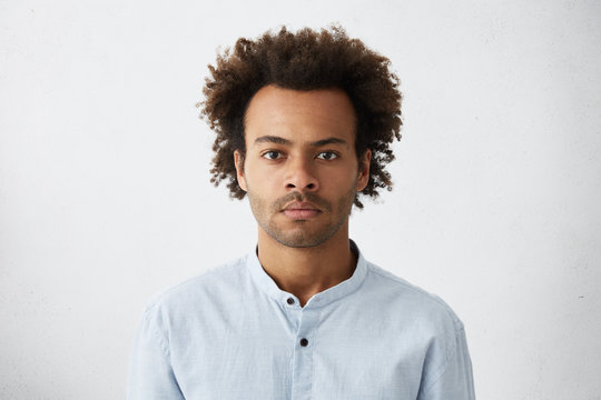 Thinking black Afro American man with serious expression looking with dark narrow eyes directly into camera while posing against white studio wall. Grave businessman in shirt thinking about work