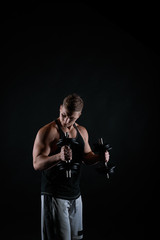 Fototapeta na wymiar Dark concept of hard training on black background. Athletic brutal man working with dumbbells. Bodybuilder trainer in t-shirt and grey shorts. Muscular guy making exercises.