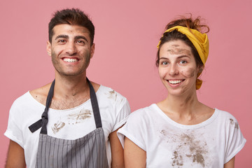 Studio portrait of smiling man and woman in white T-shirts being dirty after cleaning their apartment isolated over pink background. Houseowners with glad experssions looking directly into camera