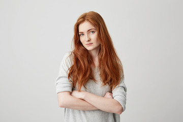 Portrait of beautiful ginger girl with freckles looking at camera with crossed arms over white...