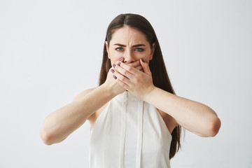 Displeased young brunette girl covering mouth with hand looking at camera over white backround.