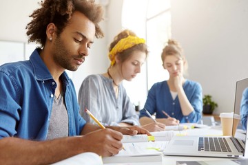 Three multiethnic people sitting in light room at desk working with papers having takeaway coffee and laptop on table. Busy mixed race students preparing for exams using different literature, internet
