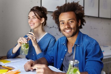 Dark-skinned male with bushy hairstyle and bristle smiling while sitting at desk near his groupmate who is drinking lemon compote working with books. Creative mixed race workers working together