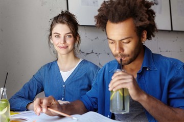 Portrait of handsome dark-skinned male drinking lemon lemonade holding pen having concentrated look into book and his female friend with beautiful appearance. Diversity, friendship, education concept