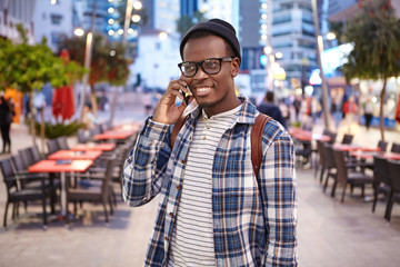 People, communication, travelling and modern technologies concept. Pleasant-looking male in checkered shirt and big eyeglasses smiling while having conversation with his friend standing in big city