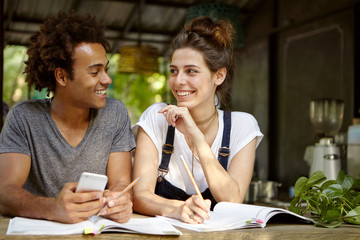 Cheerful young people of different races studying in coffee shop using books and mobile phone looking at each other with smiles. Couple in love being busy with their project work or course paper