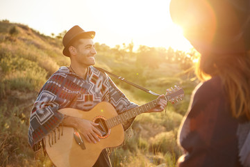 Cheerful talented male playing guitar enjoying his hobby standing near his lover demonsrtating his talent to her. Man in love singing songs proposing his girlfriend to marry in romantic manner - obrazy, fototapety, plakaty