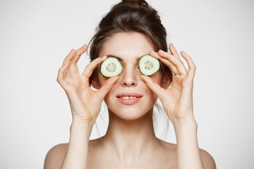 Young beautiful nude girl smiling hiding eyes behind cucumber slices over white background. Beauty skincare and cosmetology.