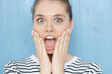 Cute young caucasian female in navy striped t-shirt with opening mouth widely, saying omg, wow, having excited astonished look, holding hands on her face standing against on blue background