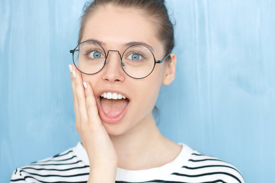 I can't believe this! Amazed young european female wearing nerdy round glasses and striped t-shirt holding hand on her face in surprised gesture, keeping mouth wide open, looking shocked and amazed