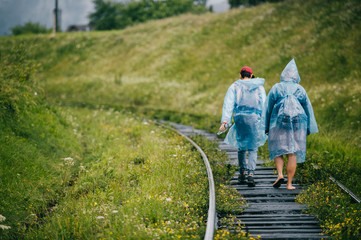 Couple of young rebel lovers travelers in raincoats walking in danger zone on railroad outdoor at nature. Company of friends rest on vacation. Tourist clothes. Hipster lifestyle. Warning. Occurence.