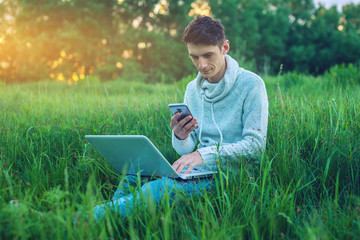 Young man sitting on a green meadow with laptop and using the phone on the background of blue cloudy sky at sunset