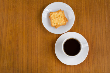 Black coffee in white ceramic cup on saucer over dark teak wood background served with cream crackers on white plate with copy space for text insertion, top view (flat lay)