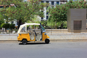 Tuk tuk small passenger three weel mini car isolated on summer empty street road background. Bright yellow rickshaw driven by locals helps tourists to travel around the city fast and cheaper than taxi