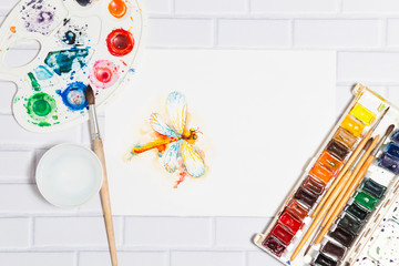 Hand Drawn Sketch of Watercolor Orange Dragonfly,with lying paints, paintbrushes and palette on the white brick background - concept of human creativity,top flat view.Have an empty place for your text