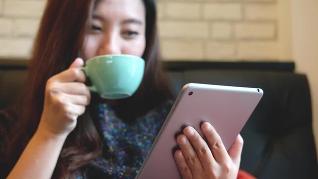 A beautiful Asian woman with smiley face holding and using tablet pc while drinking hot coffee in modern cafe 