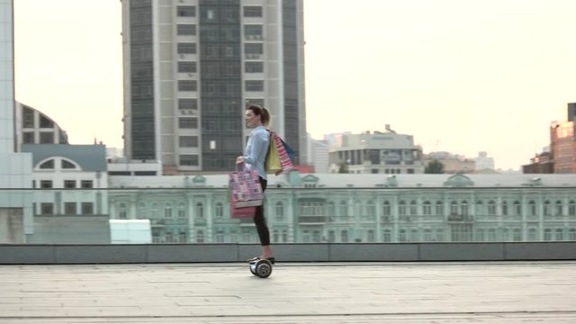 Woman riding hoverboard, shopping bags. Young female on city background. Buy and spend.