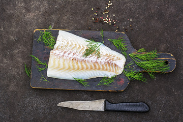 Raw cod before cooking on a black chopping Board with herbs and sea salt on a dark background. Top view