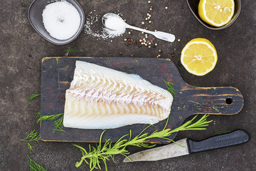 Raw cod before cooking on a black chopping Board with herbs and sea salt on a dark background. Top...