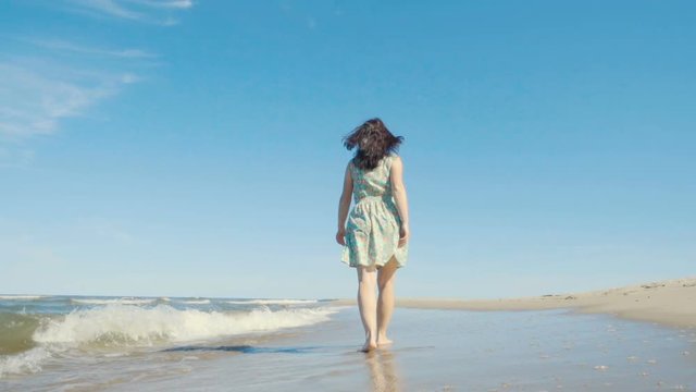 Attractive young woman in a dress strolls along the seacoast barefoot and enjoys herself. Beach with white sand. Outgoing coastal line. Slow Motion.