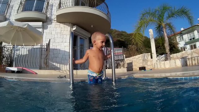 Bald toddler stands and plays on the ledder to the swimming pool as viewed from the water side