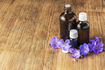 Essential oil of geranium meadow in dark glass containers on wooden background with flowers and leaves. Selective focus.