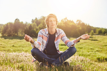 Outdoor portrait of teenger sitting on green grass crossed hands closing his eyes meditating trying to relax and to concentrate enjoying calm atmosphere of nature. People, meditation, enjoyment