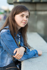 27 year old girl without makeup with freckles in a jeans jacket stands near a stone fence and watches in the distance