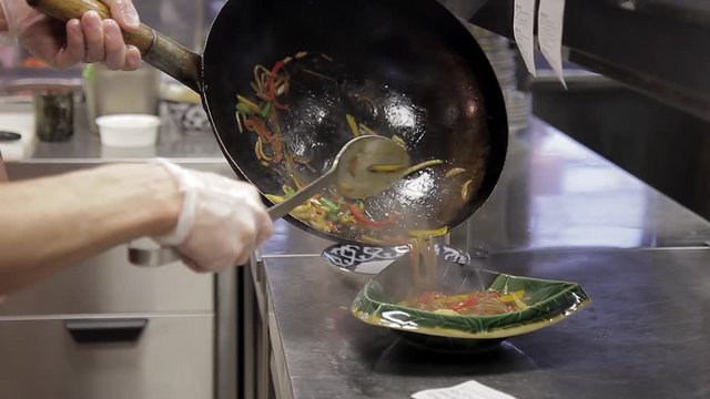 male chef put Chinese noodles with shrimps with spicy vegetable sauce from fry pan to black deep dish, dressed in dark uniform decorating pasta salad in industrial kitchen. Chef decorating plate and