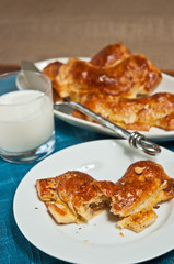 Opened coconut, almond bear claw, danish ;pastry on a white, round plate and a whole bear claw on an other plate with an artisan spoon, a glass of milk all on a blue place mat