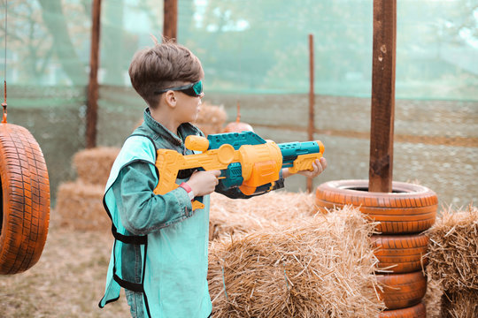 Young boy with blaster attack and play with friends in protective glasses. Excited Child with darts toy gun on the play field