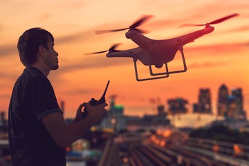 Silhouette of man controlling a drone at sunset. City in background. - obrazy, fototapety, plakaty