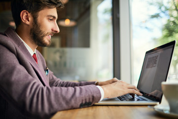 Side view of young man typing documents on laptop while in a cafe