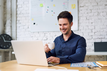 Fototapeta na wymiar Joyful young startup businessman enjoying coffee and working on laptop in modern home office
