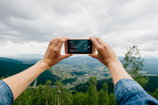Closeup of tan women hands holding mobile phone with wide touchscreen and making photos and video of beautiful nature landscape from high altitude in mountains.  Low cloudy sky. Travel and dicover.