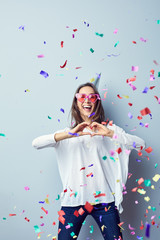 Laughing young woman dressed in heart shaped glasses making hear gesture at camera with confetti around her