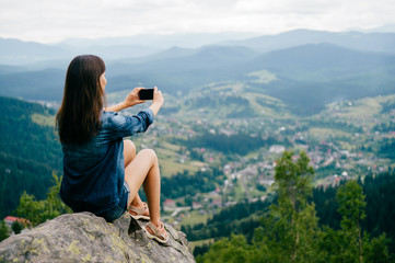 Naklejka premium Young and beautiful skinny long haired girl making photos on phone cam of nature landscape from high altitude at top of mountain with excellent sightseeing view. Travel, trip, vacation, weekend voyage