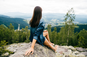 Naklejka na ściany i meble Lonely young traveler girl siiting on stone at top of mountain. Discovering new country. Beautiful nature landscape from high altitude. Summer trip on weekend vacation. Edge of world. Jeans clothes.