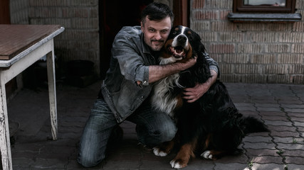 Man in an embrace with a dog (breed of mountain dog) looking into the camera against the backdrop...