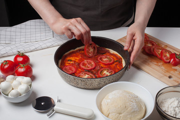 Female chef adds tomatoes to make homemade pizza. On the white table are mozzarella, pizza dough
