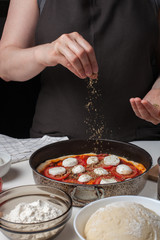 Hands of the woman cook sprinkle Italian raw pizza margarita oreano on a dark background. On the white table lie the ingredients of mozzarella, garlic, yeast dough, flour and olive oil