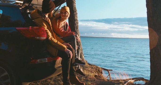 DOLLY IN Young Caucasian couple sitting on the trunk of a modern SUV and enjoying the view of a sunset over the large lake. 4K UHD 60 FPS slow motion