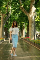 Young woman is walking in a park in a white blouse and a blue pencil skirt.