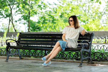 Young woman in a park in a white blouse and blue skirt is sitting on a bench.