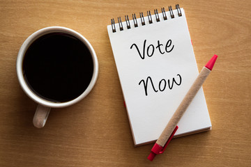 Vote now handwriting on notebook with cup of coffee.