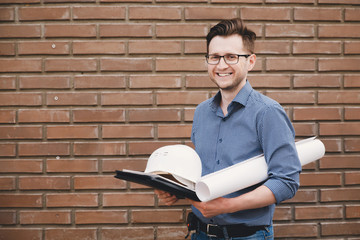 man builder with glasses smiles and stands on the background of a brick wall, a helmet, a folder, drawings are in hands. Concept building of high-rise buildings, businessman, foreman.